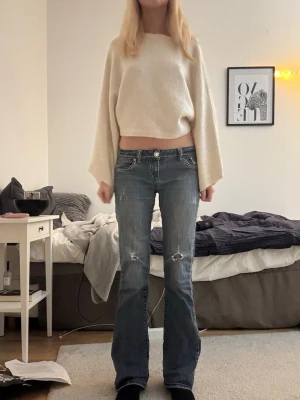 Blå bootcut jeans med slitningar - Säljer ett par blå bootcut jeans från LA idol med slitningar på låren och snygga broderade detaljer på bakfickorna. Jeansen har låg midja, vita sömmar och dekorativa knappar.