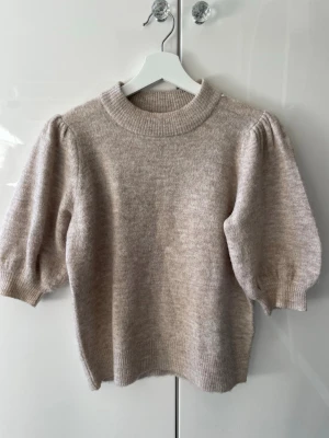 Beige puffärmstickad tröja ONLY - Supersöt beige stickad tröja från ONLY med puffärmar och ribbade muddar. Tröjan har en rund halsringning och är gjord i ett mjukt, mysigt material som känns lyxigt. Perfekt för dig som gillar trendiga detaljer och en clean look.