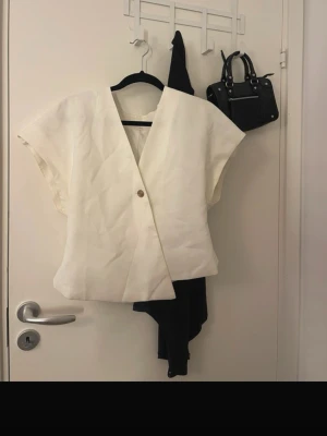 Vit omlottopp med kort ärm - Stilren vit omlottopp med v-ringning och korta ärmar. Toppen har en enkel knapp framtill och är tillverkad i ett lätt tyg som ger en luftig känsla. Perfekt för en trendig och avslappnad look.