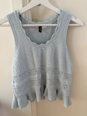 Ljusblå stickad linnetopp med volang - Supersöt ljusblå stickad linnetopp från Gina Tricot med volangkant nertill och vacker hålstickad detalj. Toppen är ärmlös och har en scallopad kant vid halsen. Perfekt att styla med jeans eller kjol för en trendig look🩵