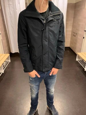  hollister vindjacka  - Snygg svart vindjacka från hollister  med hög ribbad krage och dragkedja framtill. Jackan har en stilren design och är perfekt för dig som vill ha en enkel men cool look. Passar bra till jeans och sneakers. Jackan har långa ärmar och diskreta detaljer vid kragen.