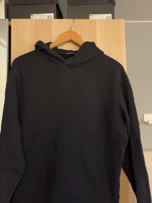 Svart hoodie från Acne Studios - Säljer en svart hoodie från Acne Studios med det klassiska face-logotypen på bröstet. Hoodien har huva och en stor magficka framtill. Tillverkad i mjuk bomull och har en avslappnad passform, perfekt för en chill och stilren look.