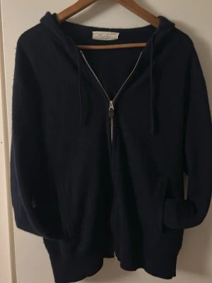 Kashmir hoodie från Sayless - Säljer en stilren mörkblå Kashmir hoodie med dragkedja och huva från Sayless. Hoodien har en klassisk passform och är perfekt för en avslappnad look. Materialet känns mjukt och bekvämt, och den har fickor framtill. Perfekt för dig som gillar enkel och tidlös stil.