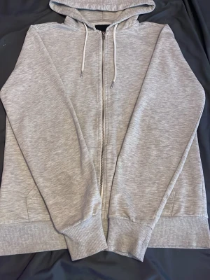 Ljusgrå zip-hoodie från 157 - Ljusgrå hoodie med dragkedja från 157. Mjuk insida, huva med vita snören och två fickor framtill. Perfekt för en avslappnad stil och enkel att matcha med andra plagg. Ribbstickade muddar vid ärmslut och nederkant.