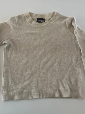 Beige stickad tröja från Pieces - En stilren beige stickad tröja från Pieces med rund halsringning och ribbade muddar vid ärmslut och nederkant. Perfekt att matcha med jeans eller kjol för en enkel och snygg look. Lite utöjd pga användning men fortfarande fin