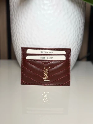 Vinrött kortfodral från Saint Laurent - Snyggt vinrött kortfodral i läder från Saint Laurent med guldfärgad YSL-logga framtill. Fodralet har flera kortfack och stilrena sömmar i matchande färg. Perfekt för att hålla koll på dina kort med stil.