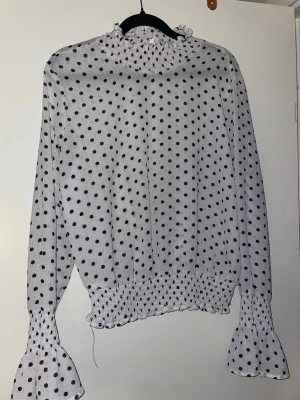 Vit prickig blus med volangärm - Trendig vit blus med svarta prickar och höga volangärmar. Blusen har hög krage med rynk och smockdetaljer vid midja och ärmslut. Perfekt för dig som gillar en lekfull och chic stil.