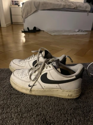 Nike Air Force 1 vita med svart swoosh - Klassiska Nike Air Force 1 sneakers i vitt läder med svart swoosh-logga på sidorna. Skorna har perforerad tå, platt sula och lågt skaft. Ikonisk streetstil med snörning och diskret Air-detalj på sulan. Perfekta för dig som gillar tidlös och clean look.