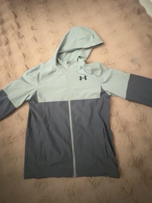 Grå vindjacka med huva från Under Armour - Snygg tvåfärgad vindjacka från Under Armour i ljusgrått och mörkgrått. Jackan har dragkedja framtill, huva och två sidofickor. Tillverkad i lätt och smidigt material som passar perfekt för blåsiga dagar. Under Armour-logga på bröstet.
