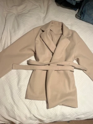Beige kappa med knytskärp - Stilren beige kappa med breda slag och knytskärp i midjan. Kappan har rak passform och är tillverkad i ett mjukt tyg som ger en clean look. 