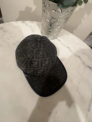 Svart keps med Fendi-logga - Pris kan vi diskutera om Snygg svart keps från Fendi med klassiskt FF-mönster över hela kepsen. Justerbar rem baktill med Fendi-logga. Kepsen har böjd skärm och är tillverkad i ett tygmaterial som känns lyxigt och trendigt.