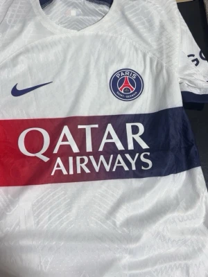 PSG vit matchtröja Nike - Snygg vit PSG matchtröja från Nike med klubbmärke på bröstet och Qatar Airways som sponsor. Tröjan har en bred röd och marinblå rand över bröstet och är tillverkad i lätt och ventilerande material, perfekt för fotboll eller träning.