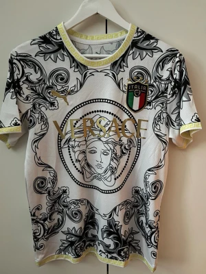  t-shirt - Säljer en vit t-shirt med barockinspirerat svart mönster och gulddetaljer. T-shirten har korta ärmar, rund halsringning med gul mönsterkant och ett stort tryck på bröstet. Materialet är polyester och designen är riktigt iögonfallande.