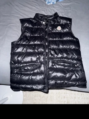 Svart dunväst från Moncler - Snygg svart dunväst från Moncler med glansig finish och klassisk logga på bröstet. Västen har ståkrage, dragkedja framtill och två fickor med dragkedja. Perfekt för lager-på-lager och streetstyle. Det går att skanna nfc