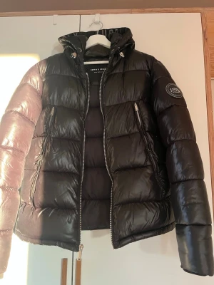 Svart pufferjacka från Supply & Demand - Svart pufferjacka från Supply & Demand med huva och två dragkedjeförsedda fickor. Jackan har en patch på ärmen och blanka paneler. 