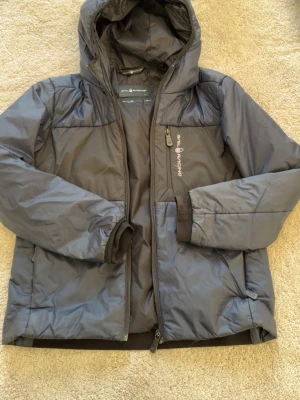 Svart pufferjacka från Sail Racing - Snygg svart pufferjacka från Sail Racing med huva och dragkedja framtill. Jackan har diskret logga på bröstet och är tillverkad i ett slitstarkt, vattenavvisande material. Perfekt för kalla dagar när du vill hålla värmen och ändå se stilren ut.