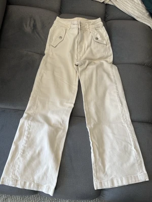 Vida beige jeans med fickdetaljer - Snygga beige jeans med vida ben och coola fickor framtill med lock och knapp. Hög midja och raka ben ger en trendig siluett. Perfekta för dig som gillar en avslappnad men stilren look. Materialet känns mjukt och skönt mot huden.