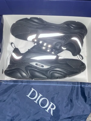 Svarta Dior B22 sneakers - Snygga svarta Dior B22 sneakers med reflekterande detaljer och chunky sula. Skorna har mesh och läderpaneler, samt tydlig Dior-logga på sidan. Perfekta för dig som vill sticka ut med en lyxig och modern look.