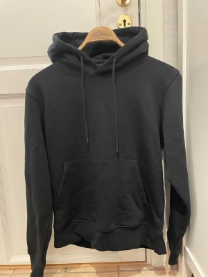 Svart hoodie från jack and jones - En klassisk svart hoodie med stor magficka och justerbar huva med snören. Hoodien har ribbade muddar vid ärmslut och nederkant. Perfekt för en avslappnad och stilren look. Materialet känns mjukt och bekvämt, troligen bomullsblandning.