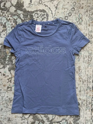 Adidas babytee  - Säljer en blå t-shirt från Adidas med stort vitt logotryck på bröstet. T-shirten har en klassisk rund hals och korta ärmar. Materialet är mjukt och bekvämt, perfekt för en avslappnad stil. Barnstorlek men passar xs/s