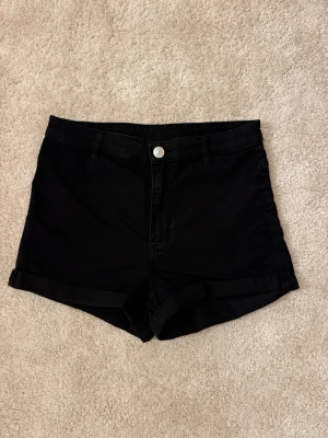 Svarta jeansshorts - Svarta shorts i jeansmaterial med hög midja och uppvikta benslut. Klassisk femficksmodell med knapp och dragkedja framtill. 