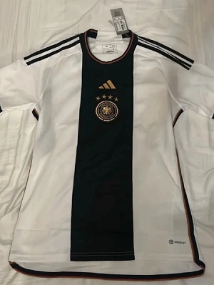 Adidas Tyskland matchtröja vit M - Säljer en vit Adidas Tyskland matchtröja i storlek medium. Tröjan har svart mittfält, guldiga detaljer och fyra stjärnor ovanför landslagsemblemet. Klassiska Adidas-ränder på axlarna och broderad logga. Perfekt för fotbollsfans!