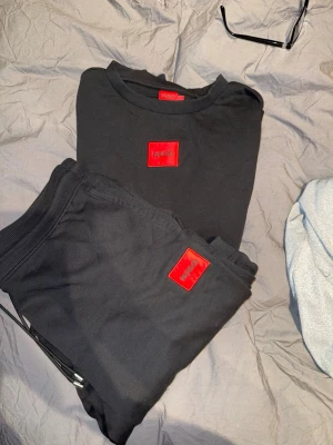 Svart mjukiset från Hugo - Snyggt svart loungewear-set från Hugo med matchande tröja och byxor. Båda plaggen har en röd patch med Hugo-loggan framtill. Mjukt material och avslappnad passform, perfekt för chill dagar hemma. Använda fåtal gånger 