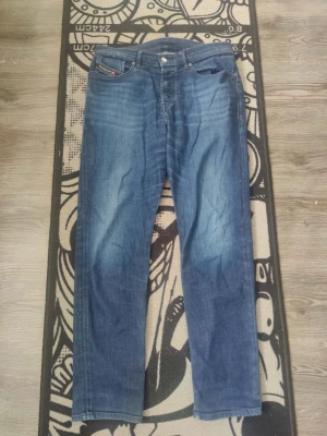 Blå raka jeans från Diesel - Snygga blå jeans från Diesel med raka ben och klassisk femficksmodell. Jeansen har en lätt tvättad look med subtila slitningar framtill och bakfickor med diskret logga. Perfekta för en avslappnad och trendig stil.