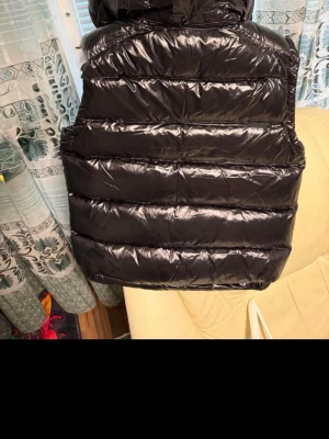 Moncler dunväst med dragkedja - Säljer en svart, glansig dunväst med hög krage och två fickor med dragkedja framtill. Västen har quiltad design och stängs med en kraftig dragkedja. Perfekt för lager-på-lager och riktigt snygg till streetwear-stil.