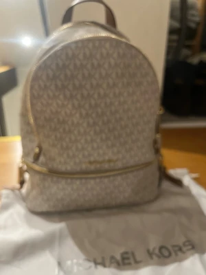 Beige ryggsäck från Michael Kors - Snygg beige ryggsäck från Michael Kors med MK-logga över hela väskan. Väskan har guldfärgade dragkedjor, ett stort huvudfack och ett mindre ytterfack framtill. Tillverkad i tåligt konstläder och har justerbara axelband. Perfekt för dig som vill ha en trendig och praktisk väska. Äkta 