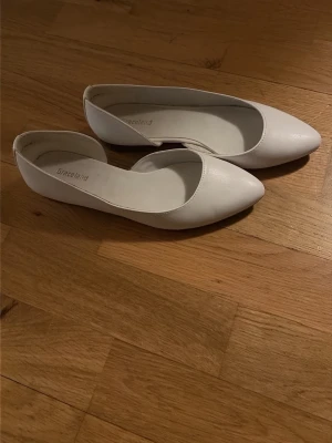 Vita ballerinaskor från Graceland - Säljer ett par stilrena vita ballerinaskor från Graceland. Skorna har spetsig tå och är gjorda i ett slätt material med låg klack. Perfekta för dig som gillar en enkel och klassisk look.