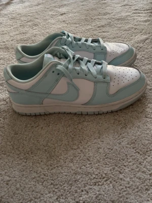 Nike Dunk Low Mint och Vit - Snygga Nike Dunk Low sneakers i mintgrönt och vitt. Skorna har klassisk låg siluett, perforerad tåbox och ikoniska swoosh-loggan på sidan. Tillverkade i läder med matchande mintgröna skosnören och slitstark yttersula. Perfekta för dig som vill sticka ut med stilrena färger.