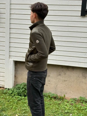 Olivgrön jacka från Moncler - Snygg olivgrön jacka från Moncler med hög krage och blandning av stickat tyg och quiltade partier. Jackan har Moncler-logga på ärmen och dragkedja framtill. Perfekt för dig som vill ha en stilren och modern look med exklusiv känsla.