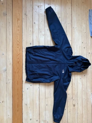 Mörkblå regnjacka från The North Face - Snygg mörkblå regnjacka från The North Face med huva och dragkedja framtill. Jackan har elastiska muddar vid ärmsluten och logga på både bröstet och ryggen. Tillverkad i vattenavvisande material, perfekt för regniga dagar. Väldigt bra skick med inga tecken på användning och riktigt skön passform!!