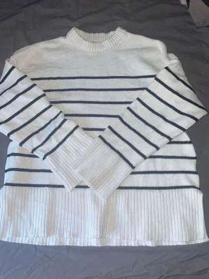 Vit stickad tröja med svarta ränder - Snygg vit stickad tröja med svarta horisontella ränder och ribbstickade muddar samt nederkant. Tröjan har rund halsringning och långa ärmar, perfekt för en stilren och avslappnad look.