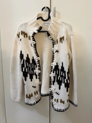 Vit och brun stickad kofta Jet Mode - Säljer en chunky stickad kofta från Jet Mode i vitt med bruna och svarta mönsterdetaljer. Koftan har dekorativa knytningar framtill, hög krage och kontrastfärgade kanter. Perfekt för dig som gillar retrovibbar och unik design.
