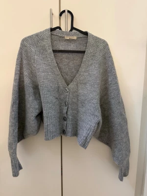 Grå ribbstickad kofta från House of Dagmar - Grå, ribbstickad kofta från House of Dagmar med v-ringning och tre knappar framtill. Koftan är croppad med långa, vida ärmar och breda muddar. Perfekt för lager-på-lager och enkel att styla till olika outfits.