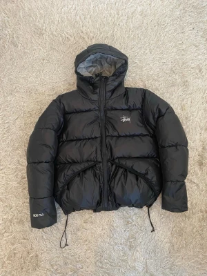 Svart pufferjacka från Stüssy - Snygg svart pufferjacka från Stüssy med huva, dragkedja och två stora fickor framtill. Jackan har Stüssy-logga på bröstet och 800 fill-brodyr på ärmen. Perfekt för kalla dagar med sin tjocka vaddering och justerbara snören i nederkant.