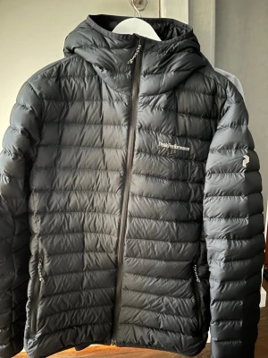 Svart dunjacka Peak Performance - Snygg svart dunjacka från Peak Performance med huva och dragkedja framtill. Jackan har två dragkedjeförsedda fickor och diskret logga på bröstet. Perfekt för kalla vinterdagar tack vare det värmande dunmaterialet och den stilrena designen.
