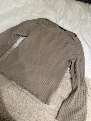 Beige långärmad topp med vågigt mönster - Trendig beige långärmad topp med vågigt strukturmönster och lätt utsvängda ärmar. Toppens material är stretchigt och ger en snygg passform. Perfekt att styla med jeans eller kjol för en cool look.