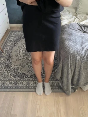 Svart kortkjol i enkel design - Säljer en svart kortkjol med rak passform och stilren look. Kjolen är enkel och klassisk, perfekt för dig som gillar minimalistiska outfits. Materialet känns mjukt och har en lätt struktur. Passar dig som vill ha en basic kjol att matcha med olika toppar. Hög midja med dragkedja.