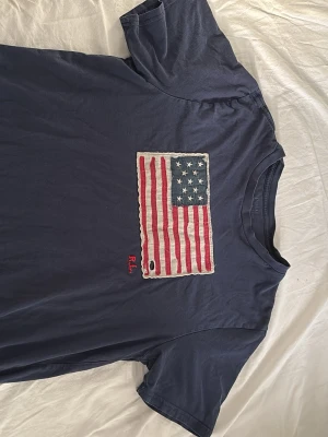 Blå Ralph Lauren t-shirt med flagga - Mörkblå t-shirt från Ralph Lauren med en stor amerikansk flagga i tyg på bröstet och RL-broderi i rött. T-shirten har klassisk passform och korta ärmar. Perfekt för dig som gillar streetwear och vill sticka ut lite extra. XL storlek men passar som en M