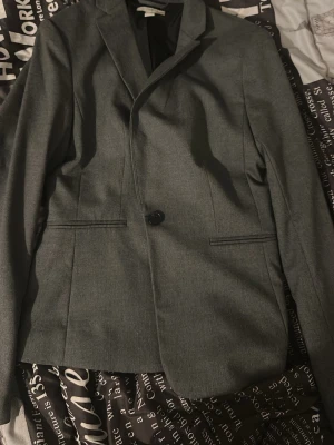 Grå kavaj med knapp - Stilren grå kavaj med en knapp framtill och två fickor. Klassisk krage och figurnära passform. Perfekt för dig som vill ha en enkel men snygg look till skolan eller jobbet.