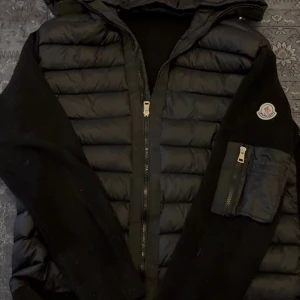 Moncler cardigan  - Snygg svart dunväst från Moncler med huva och stickade svarta ärmar. Västen har dragkedja framtill och en praktisk ficka med dragkedja på ärmen. Klassisk Moncler-logga på bröstet och quiltad design för extra värme.
