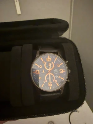 Svart klocka med orange detaljer - Snygg svart klocka med orange siffror och visare. Klockan har rund urtavla, svart läderarmband och tydliga markeringar. Perfekt accessoar för dig som gillar en tuff och stilren look.