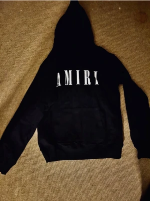 Svart AMIRI hoodie med vit logga - Svart hoodie från AMIRI med stor vit logga framtill. Tröjan har huva, känguruficka och långa ärmar. Mjuk insida och klassisk streetwear-look. Perfekt för dig som gillar exklusiva märken och stilren design. Helt ny aldrig använd