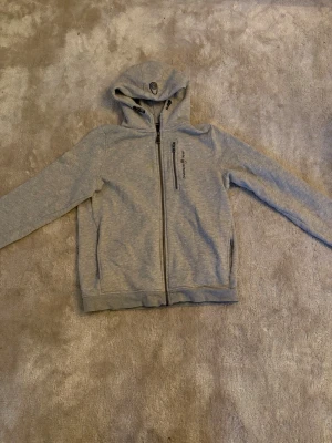 Ljusgrå zip hoodie från Didriksons - Ljusgrå hoodie med dragkedja från Didriksons. Tröjan har huva, två sidofickor och en bröstficka med dragkedja. Tillverkad i mjukt material och har en stilren look med diskret logga på bröstet. Perfekt för chill dagar eller till skolan. Storlek 168 på barn 