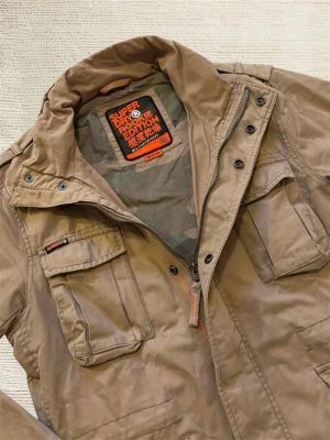 Superdry Fieldjacket - En mycket trevlig fieldjacket av märket Superdry i en extremt snygg beige färg! Hör av dig vid frågor!