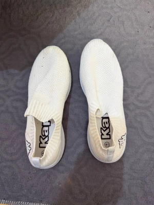 Vita slip-on sneakers från Kappa - Stilrena vita slip-on sneakers från Kappa med stickad ovandel i mesh för extra andningsförmåga. Skorna har en låg profil och elastisk mudd runt öppningen för enkel påtagning. Perfekta för en clean och sportig look.