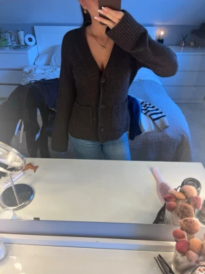 Mörkbrun kofta från Wera - Mysig mörkbrun kofta från Wera med v-ringning och stora knappar framtill. Koftan har långa ärmar, ribbade muddar och två fickor fram. Perfekt att slänga över en t-shirt eller linne för en avslappnad look.